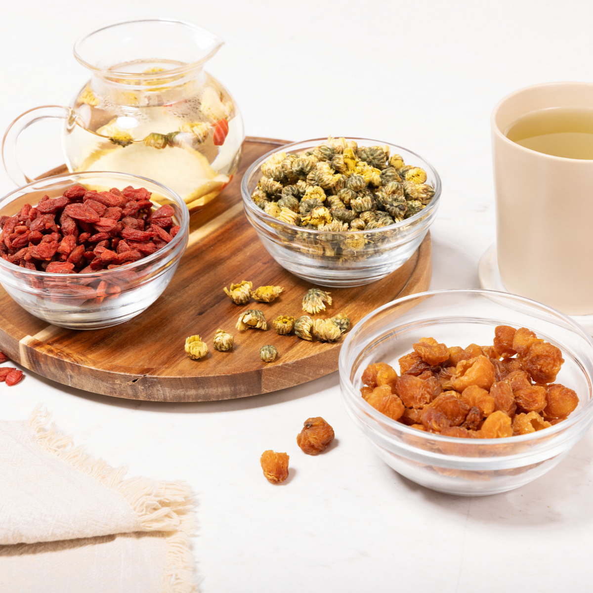A styled flat-lay of dried herbal tea ingredients on a white surface, including a glass teapot with brewed tea, bowls of Goji Berry, Baby Chrysanthemum,  and Longan Fruit