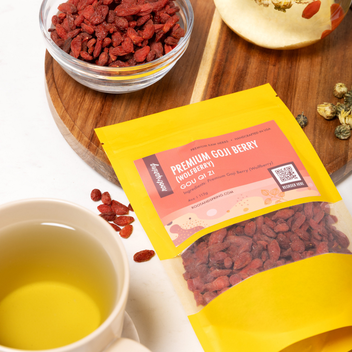 a lifestyle image of Premium Goji Berry (Wildberry) Gou Qi Pill, displayed on a wooden board next to a cup of brewed tea, a glass bowl of dried goji berries.