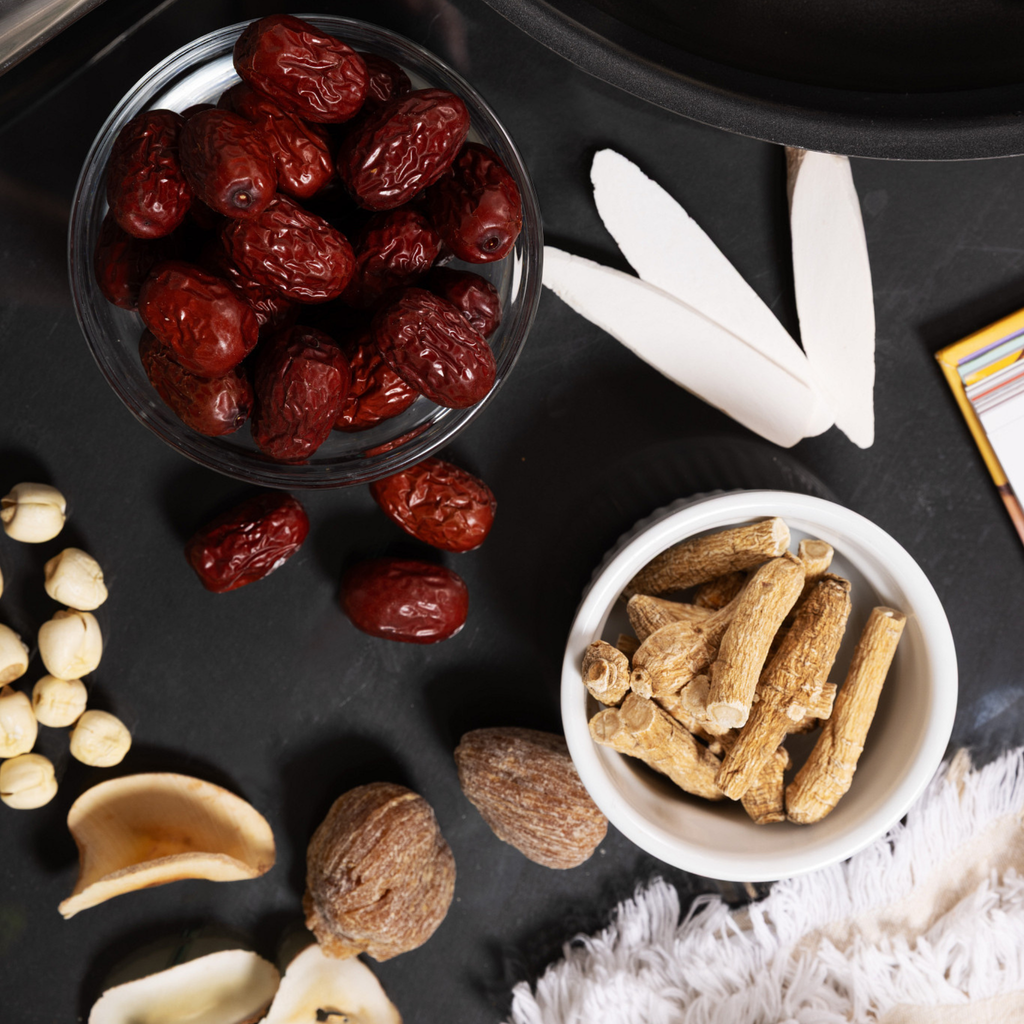 A styled flat-lay of dried herbal ingredients on a dark surface, including Red Dates, White Peony, White Lotus Seed, Candied Dates, and American Ginseng.
